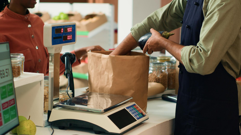 Atendente de avental colocando uma fruta em um saco de papel ao lado de uma balança digital.