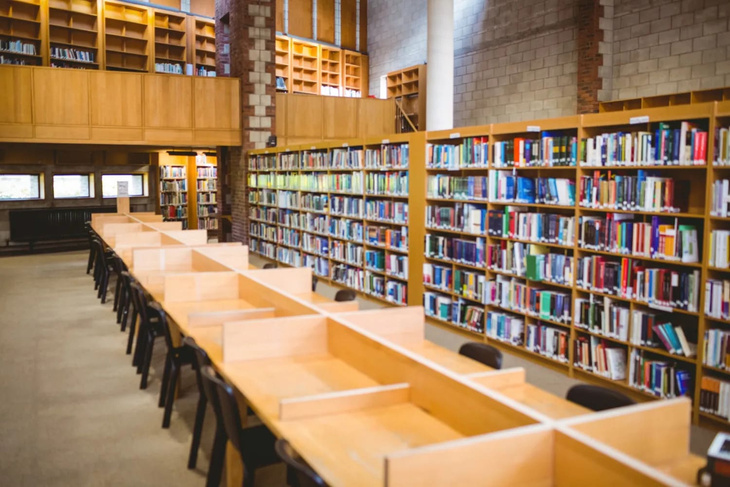 Biblioteca ampla e vazia com mesas compridas de estudo e estantes cheias de livros.