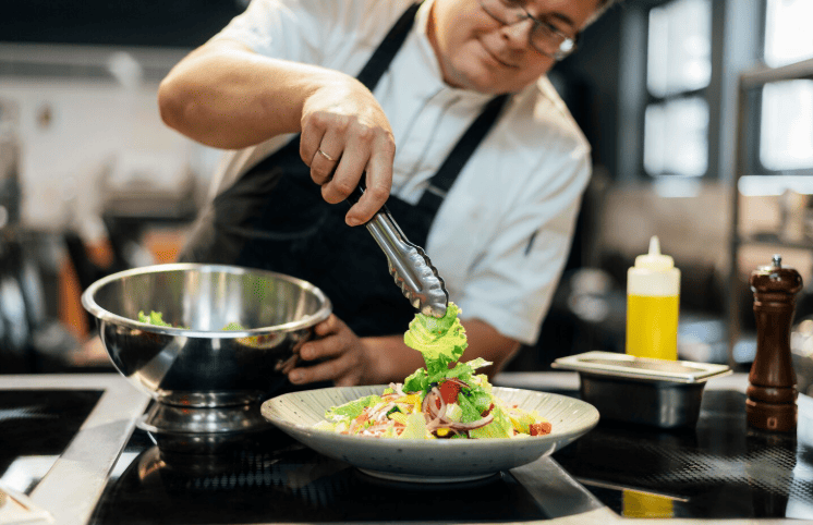 Chef de cozinha usando pinça para finalizar uma salada de alface e tomate no prato.