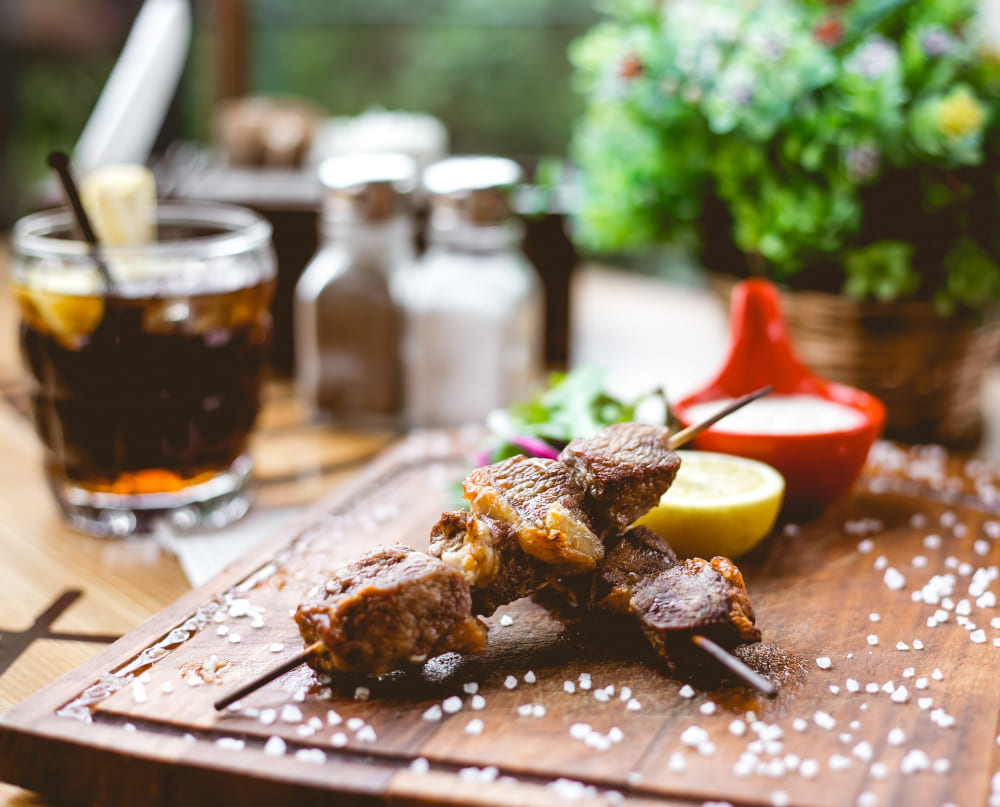 Dois espetinhos de carne grelhada servidos em uma tábua de madeira com sal grosso.