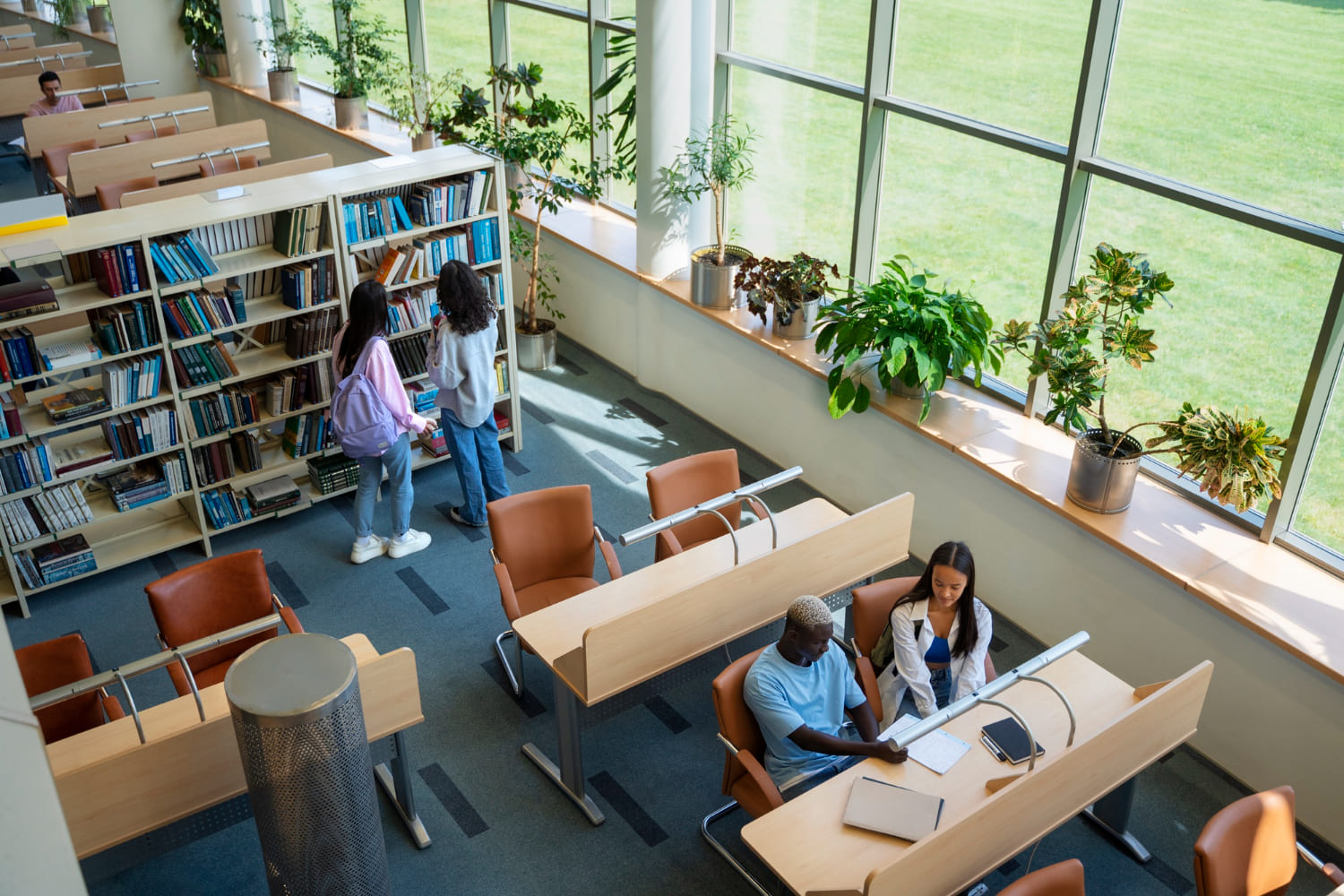 Jovens estudando em mesas ao lado de estantes de livros em uma biblioteca iluminada.
