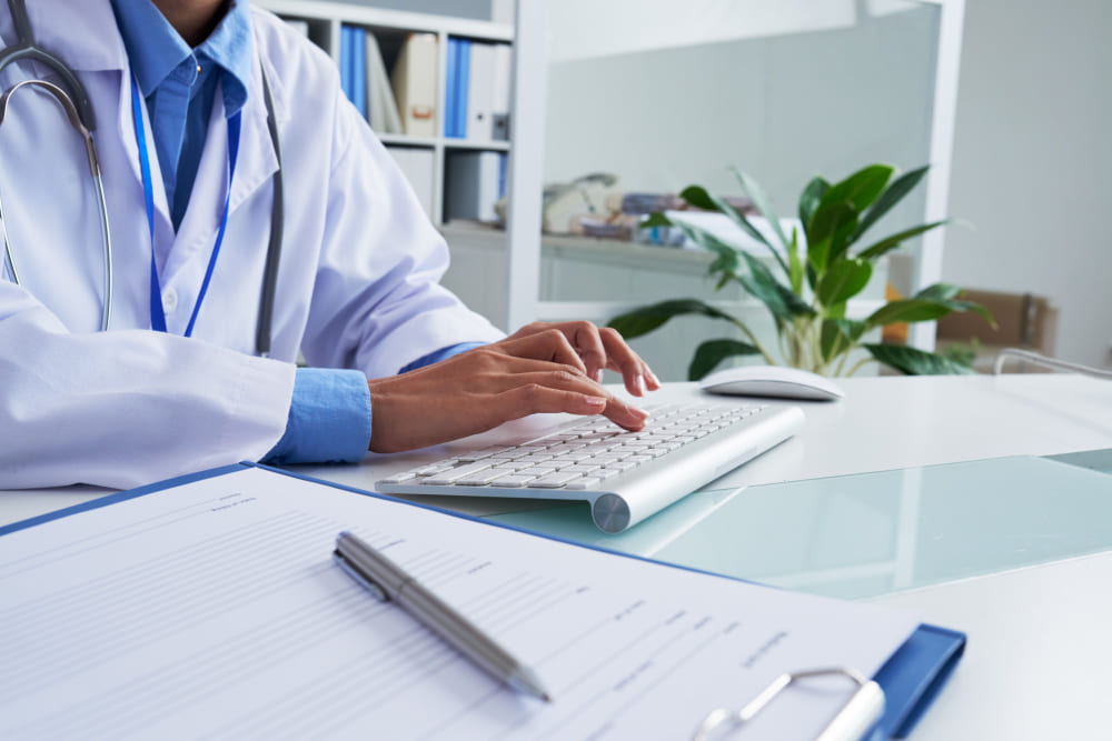 Médico de jaleco digitando no computador com prontuário sobre a mesa.