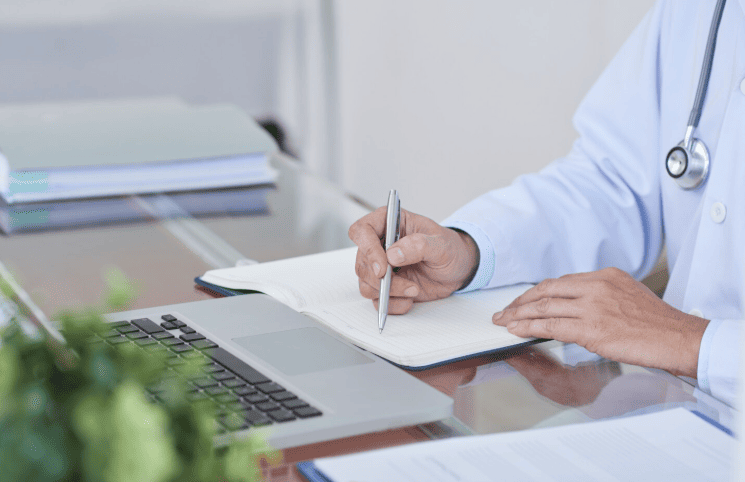 Médico de jaleco escrevendo em um caderno ao lado de um notebook.