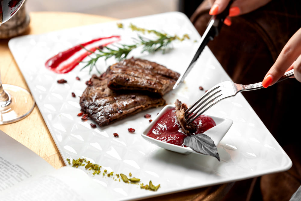  Pessoa cortando carne grelhada servida com molho vermelho em prato branco decorado.