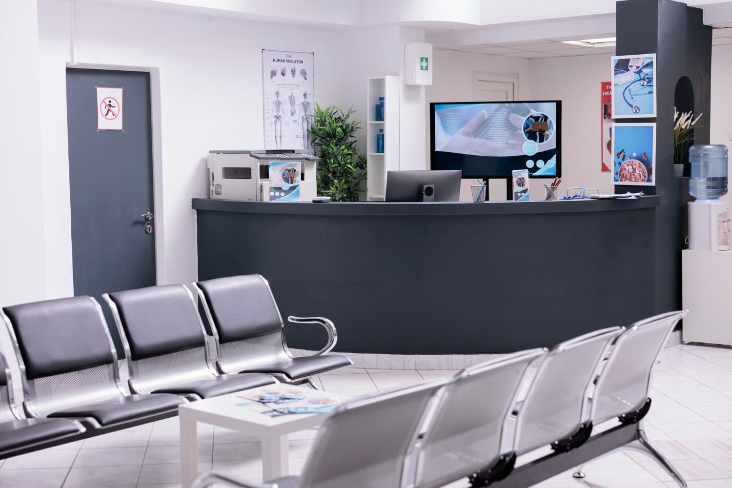 Sala de espera de uma clínica médica com balcão de recepção e cadeiras cinzas.