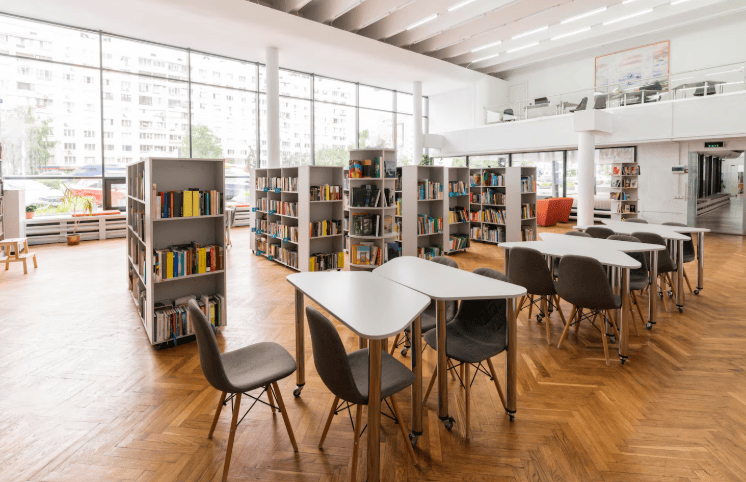 Biblioteca ampla com mesas cinzas de estudo e estantes brancas de livros ao fundo.