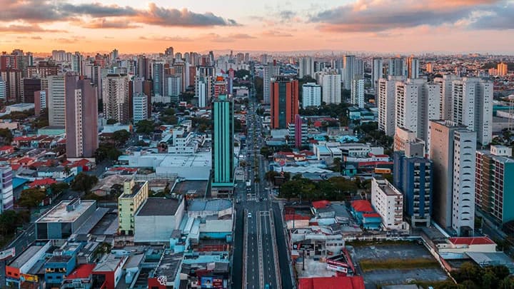 Vista aérea de uma cidade com avenida central e prédios sob o céu do pôr do sol.
